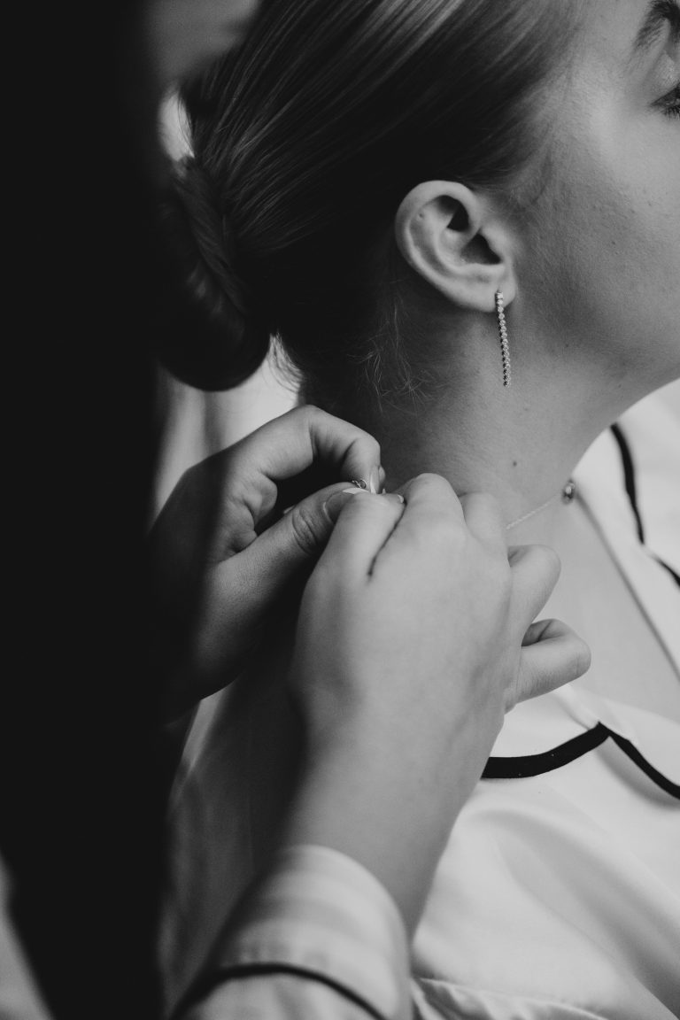 A close up photo of a bridesmaid helping the bride put her necklace on.