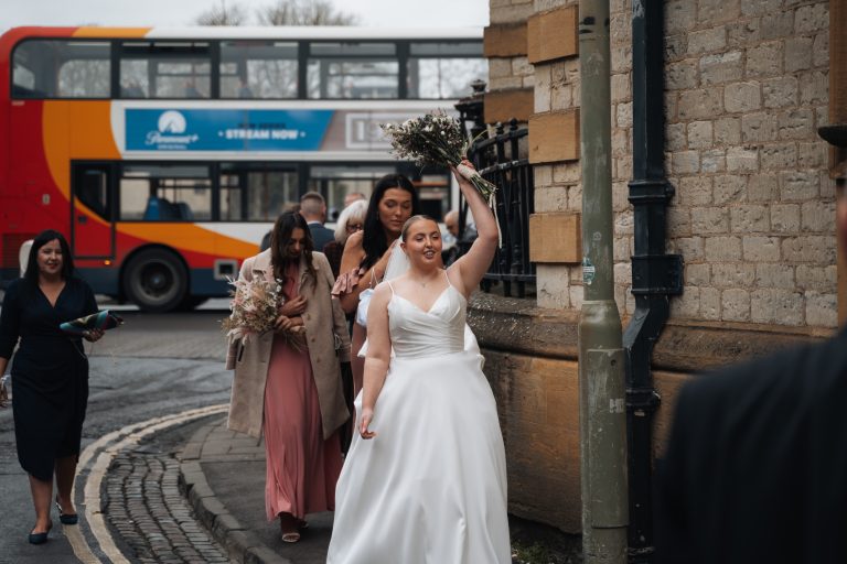 Bride and bridal party walking out of the oxford registry office with the bride holding a boquet above her head.